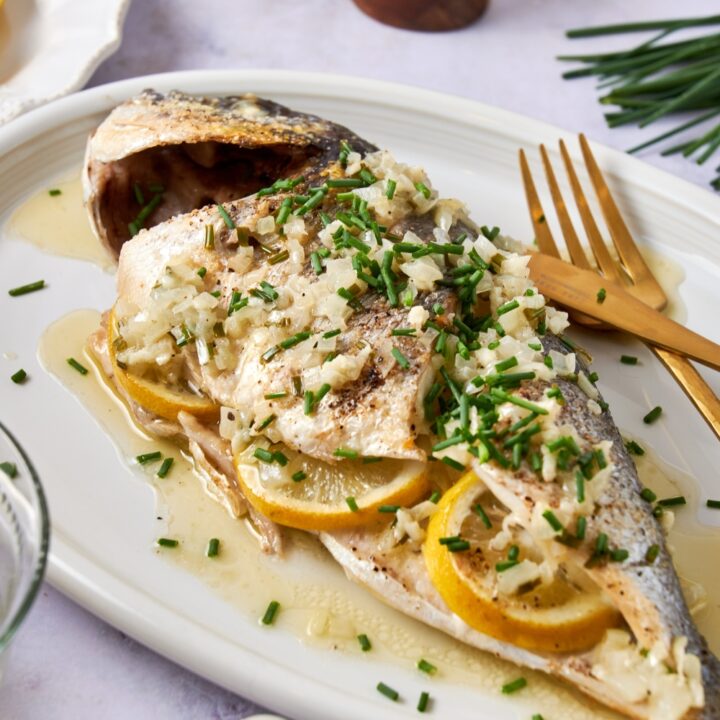 A whole cooked Branzino sits on a white platter. It is covered in lemon garlic butter sauce and stuffed with lemon slices.