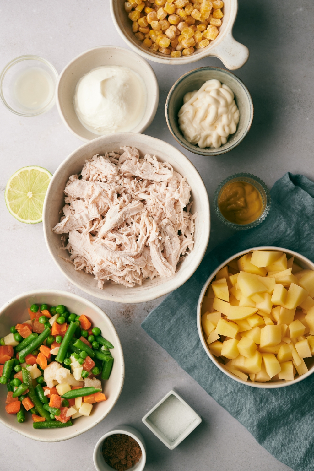 Corn, diced potatoes, mayonnaise, sour cream, shredded chicken, mixed vegetables and seasonings are in separate bowls on a white counter top.