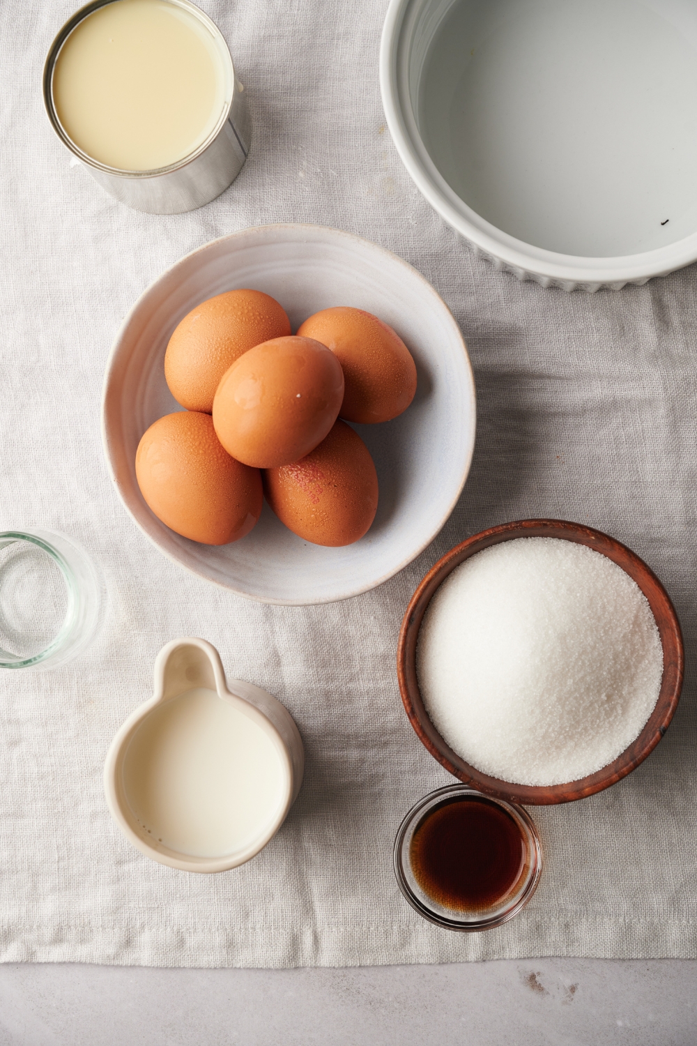 A countertop with various bowls containing ingredients like eggs, condensed milk, sugar, vanilla, milk, and water.