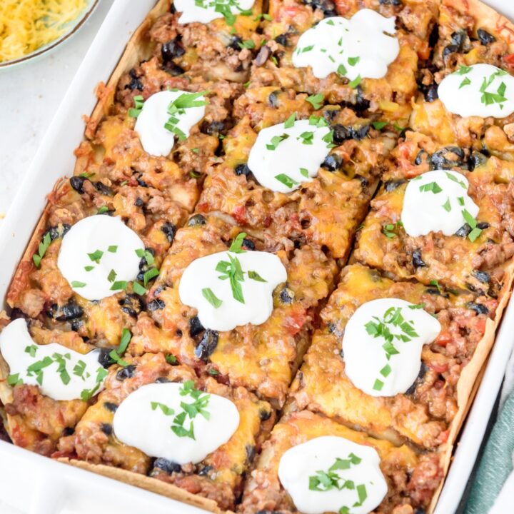 A baking dish filled with Mexican lasagna that's been cut into equal-sized pieces and topped with a dollop of sour cream on each slice.