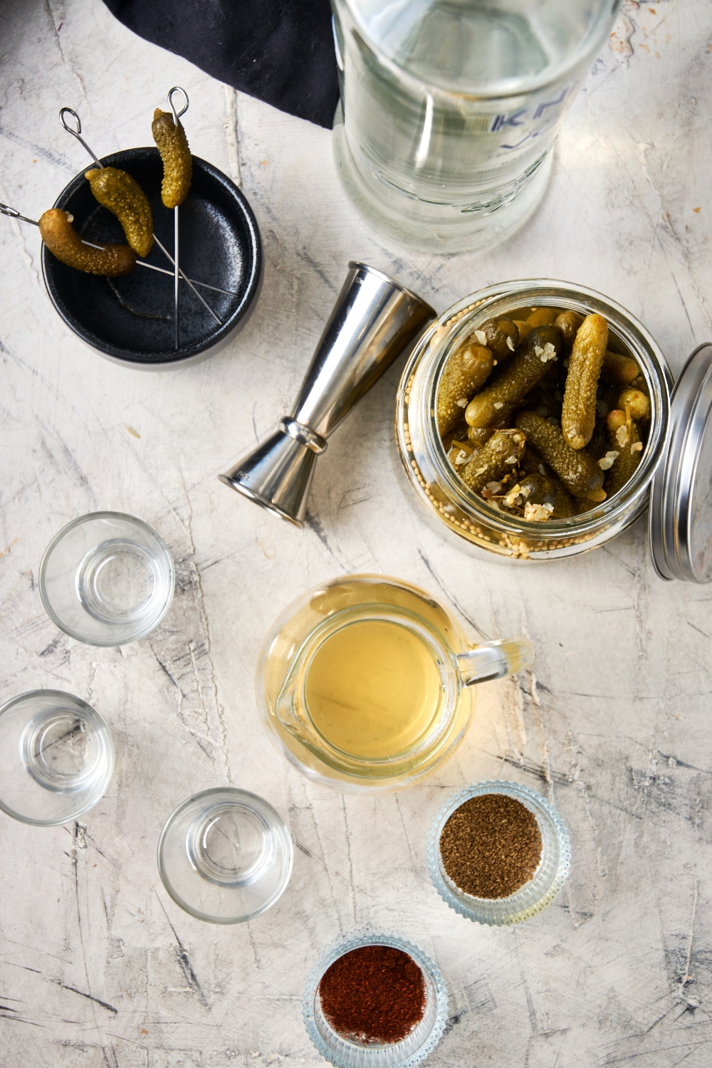 A countertop with pickle juice, tajin seasoning, pickles, vodka, and shot glasses.