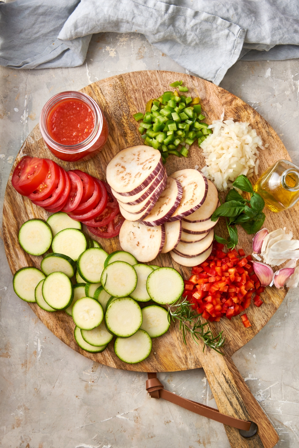 A wooden board with sliced eggplant, zucchini, tomato, diced peppers, onions, and garlic cloves are on a gray counter.