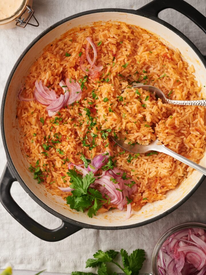 A large white pot with black handles is full of arroz rojo. Red onions and cilantro are garnishing the dish. Serving utensils are resting in the pot.