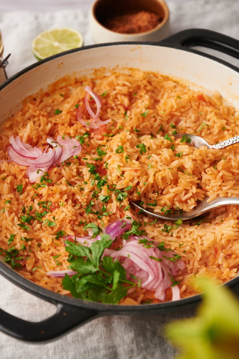 A large white pot is full of arroz rojo. Serving utensils are resting in the pot. Red onion and cilantro are garnishing the dish.