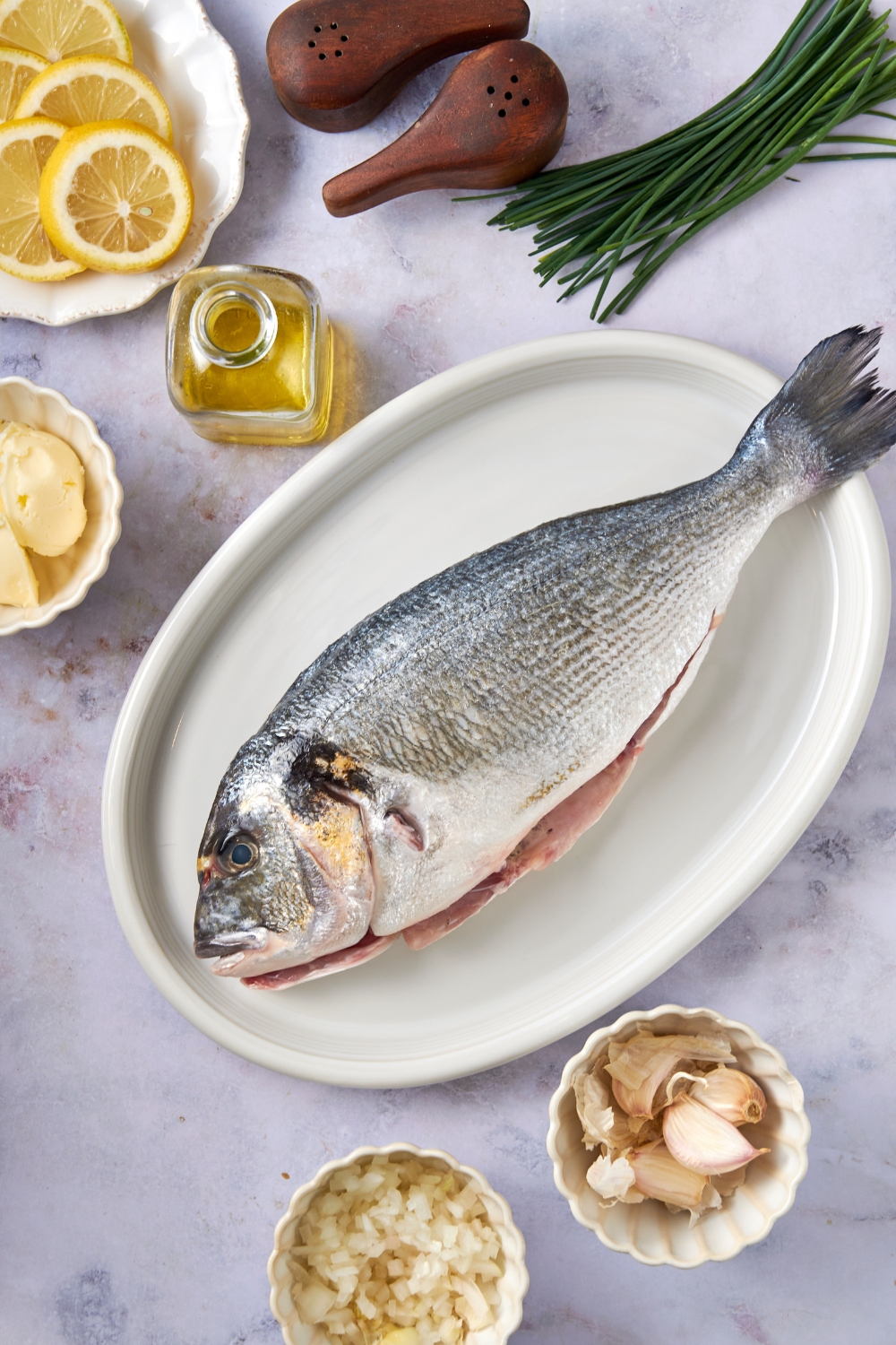 A whole Branzino sits on a white platter. Oil, lemon slices, chives, garlic and butter are in separate containers nearby.