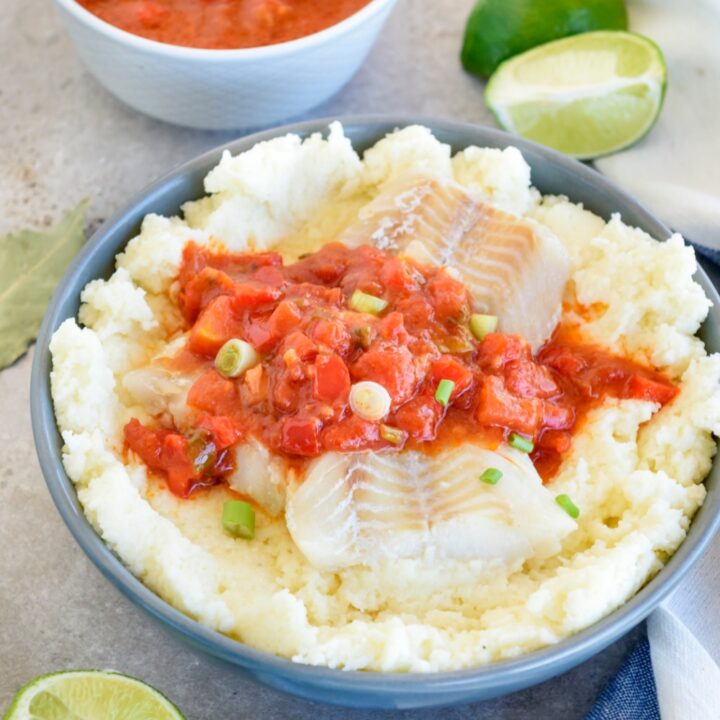 A blue bowl is full of fish and grits. Behind it, a smaller white bowl is full of tomato and vegetable sauce. Limes are nearby.