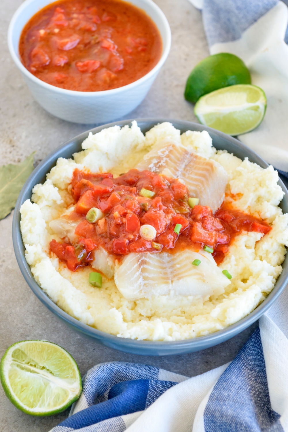 A blue bowl is full of fish and grits. Behind it, a smaller white bowl is full of tomato and vegetable sauce. Limes are nearby.