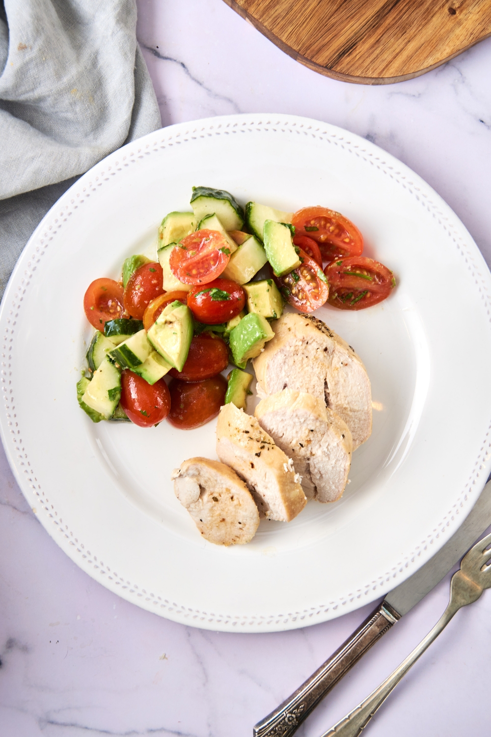 Sliced chicken breast and veggies are on a white plate. Serving utensils are beside it.