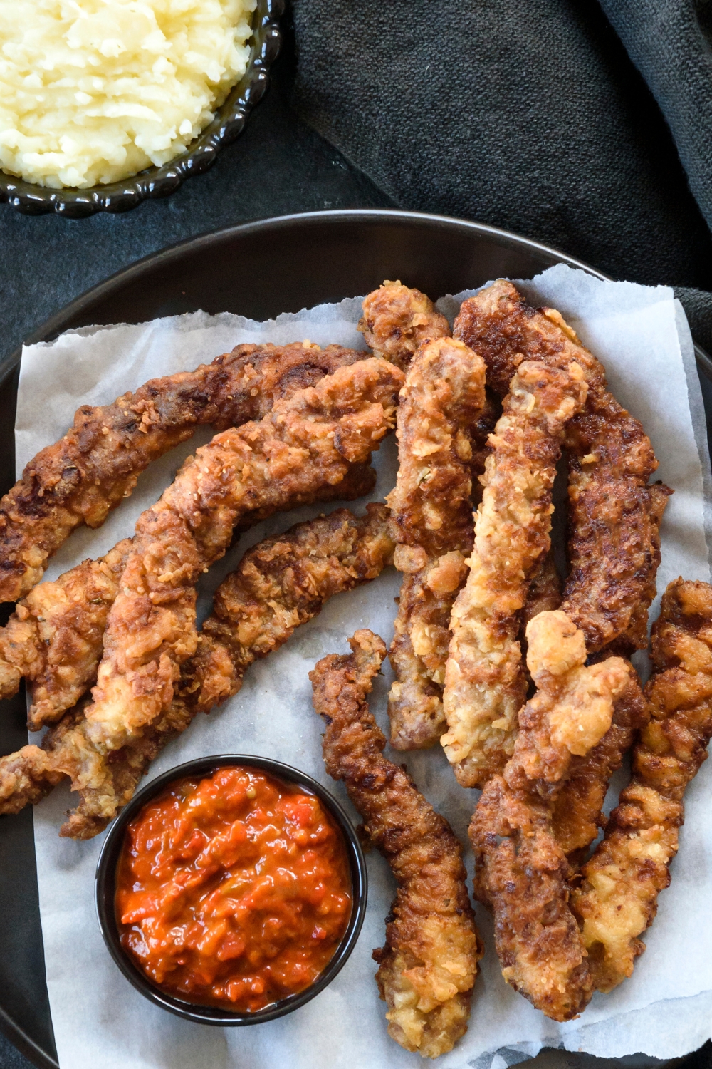 Golden brown beef cutlets sit on a paper towel lined plate. A tomato dipping sauce is in a small container and a bowl of mashed potatoes is near by.