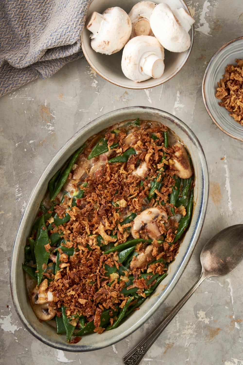A golden brown dish of green bean casserole sits on a gray counter. A bowl full of white mushrooms sits nearby.