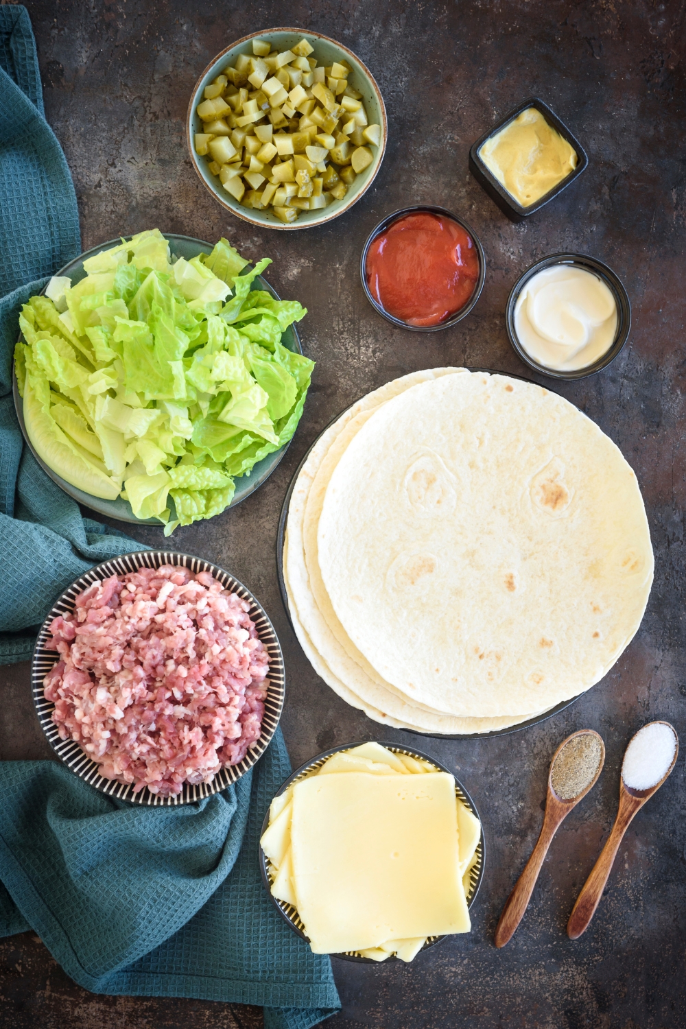 Diced pickles, condiments, shredded lettuce, ground meat, cheese, and tortillas are in separate bowls on a wooden table.