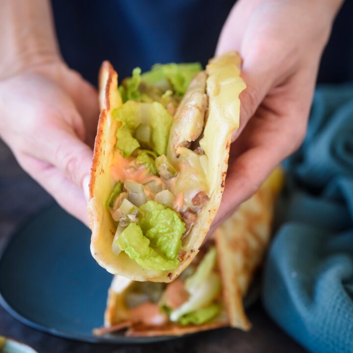 Someone holds a smash burger taco in both hands. The taco is topped with veggies and sauce.