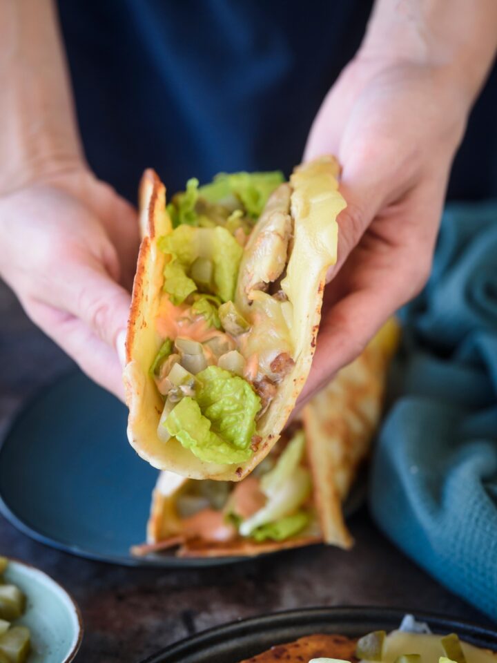 Someone holds a smash burger taco in both hands. The taco is topped with veggies and sauce.