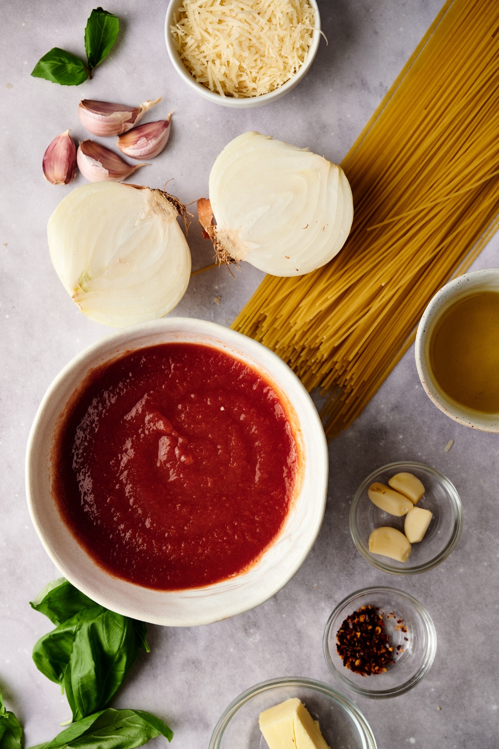 A bowl of tomato sauce, an onion, parmesan cheese, garlic, and seasonings are in separate bowls on a white counter.