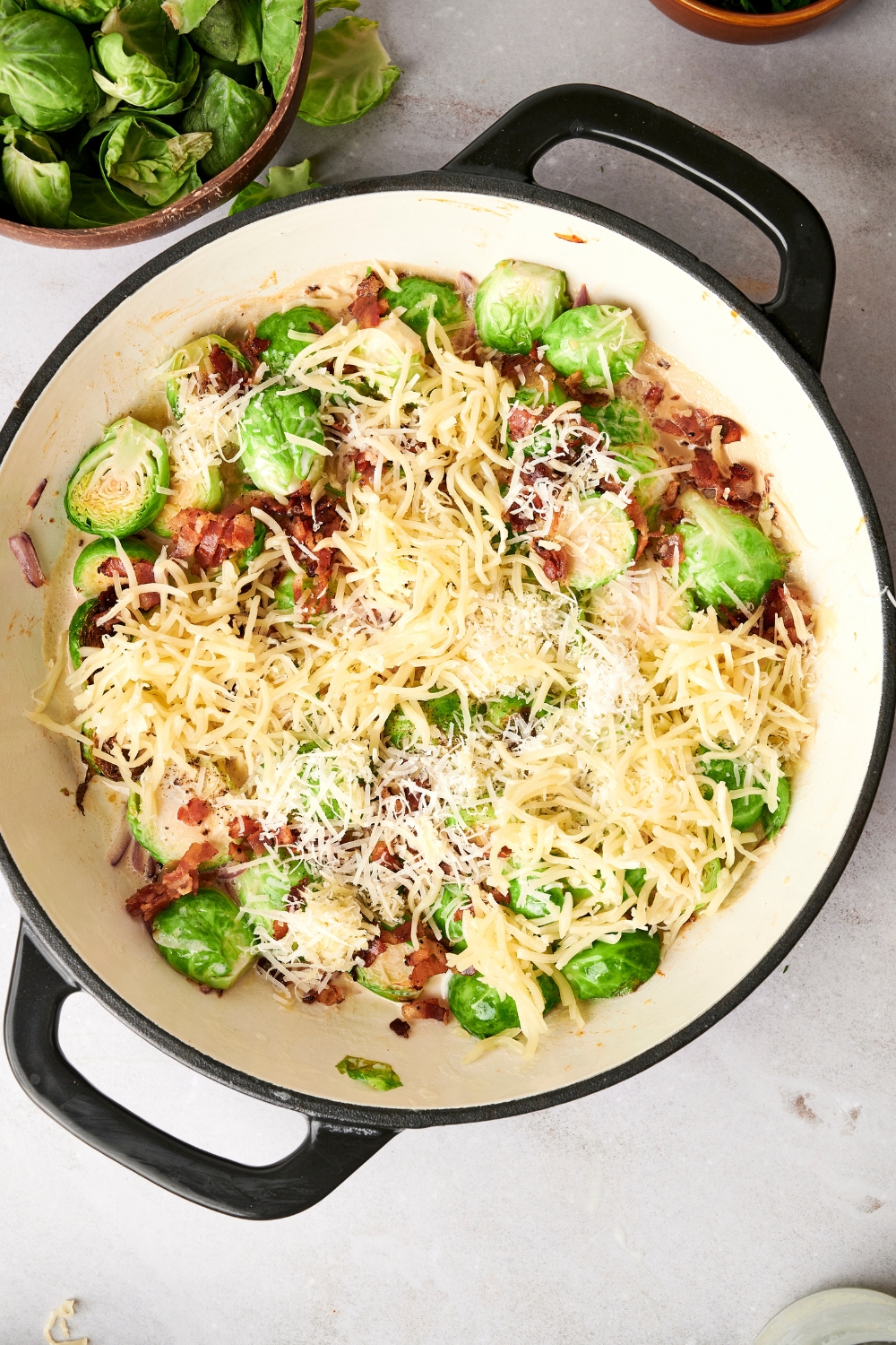 Brussel sprouts, bacon, and shredded cheese are in a white pot with black handles.