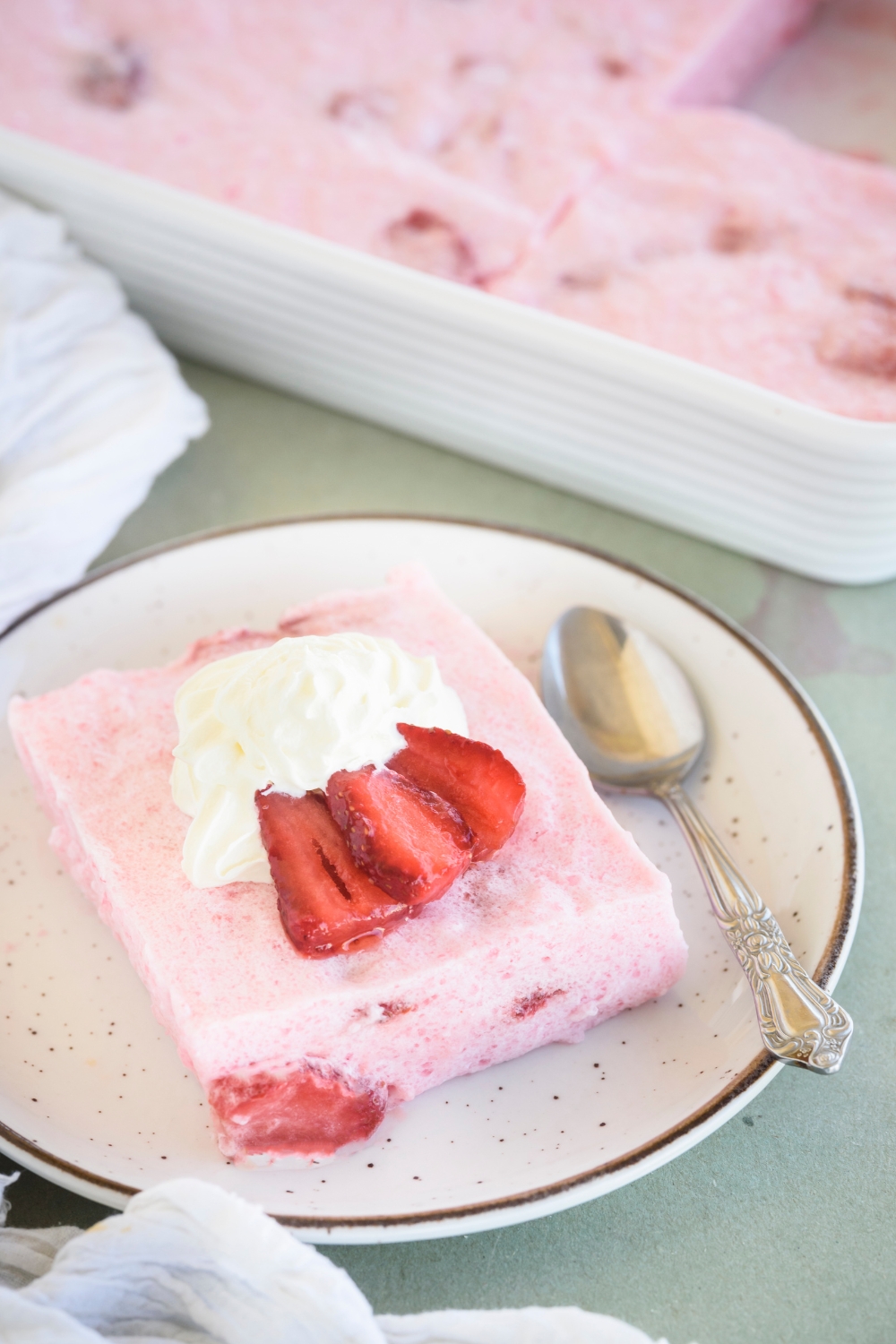A square serving of strawberry Jello fruit salad with a dollop of whipped cream and three strawberry slices on top. There is a spoon on the plate.