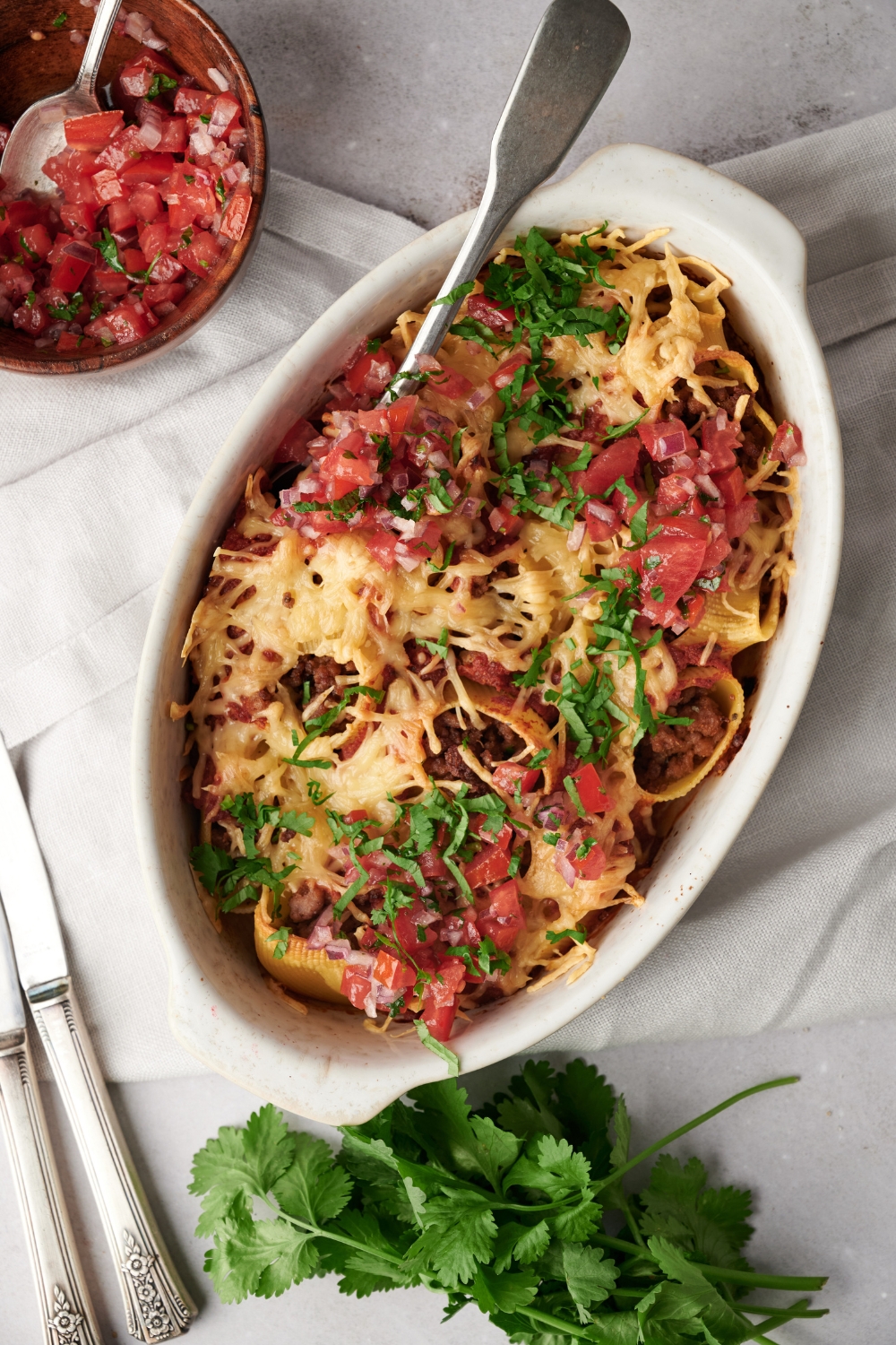 A baking dish filled with baked pasta shells covered in melted cheese, pico de gallo, and fresh green herbs.