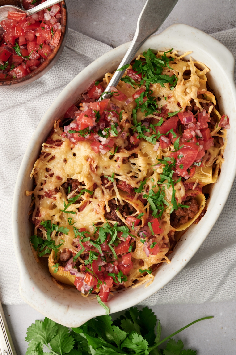 A baking dish filled with baked pasta shells covered in melted cheese, pico de gallo, and fresh green herbs.