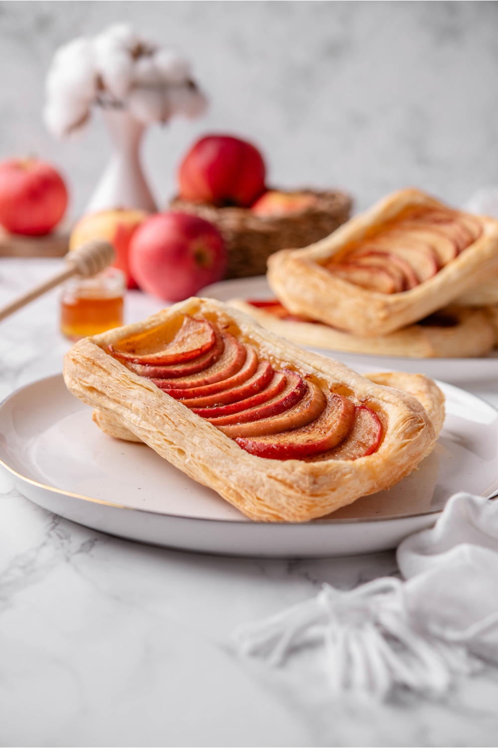 A puff pastry dough that has apples lined down the middle of it overlapping one another. The pastry is on a white plate with another pastry behind it on the plate.