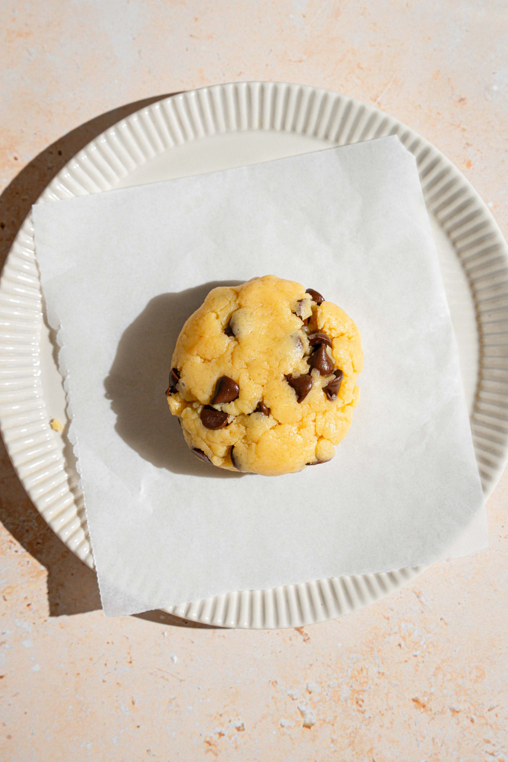 A ball of chocolate chip cookie dough on a sheet of parchment paper on a white plate.