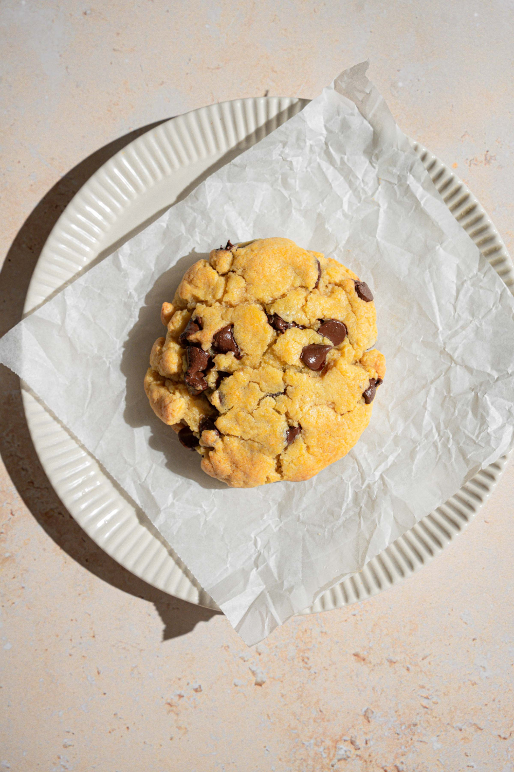 A microwaved chocolate chip cookie on top of a sheet of parchment paper on a white plate on a white counter.