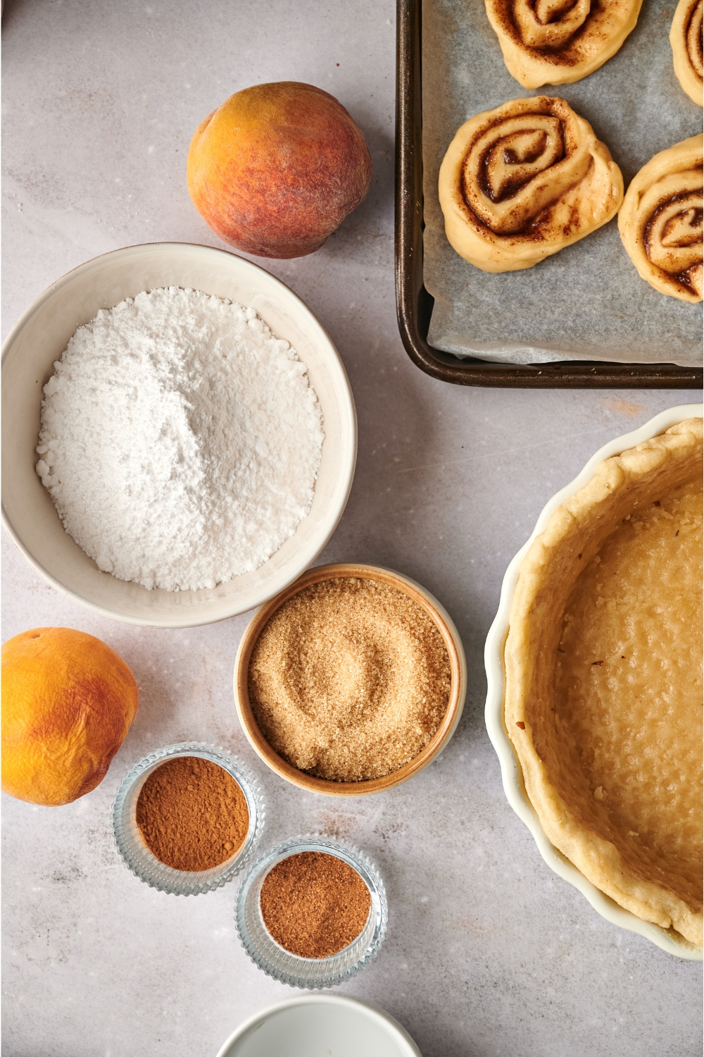 A countertop with multiple containers with brown sugar, cinnamon rolls, peaches, cinnamon, nutmeg, powdered sugar, and puff pastry.