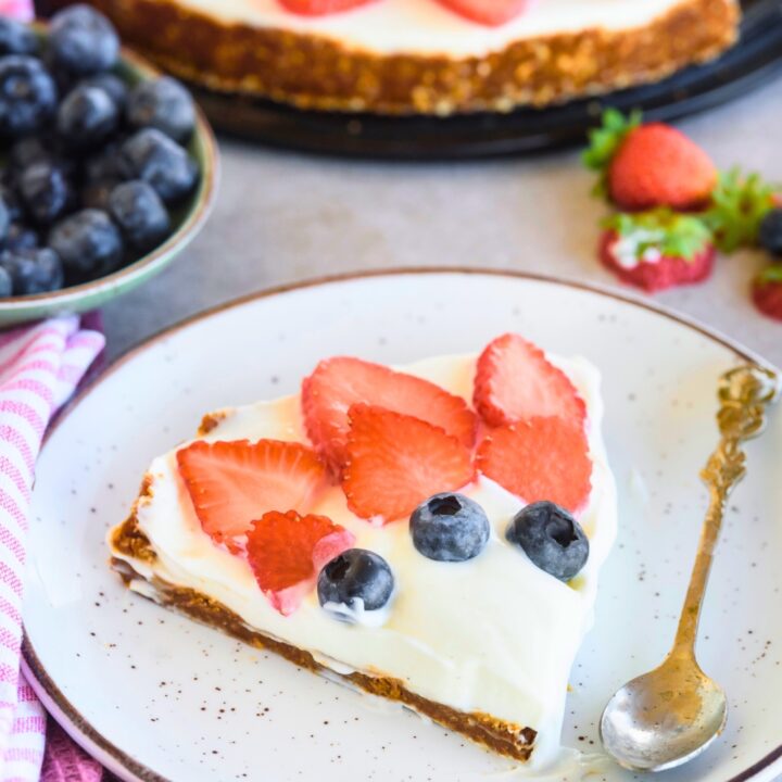 Sliced strawberries and blueberries on top of a cool whip pie on a graham cracker crust on a white plate with a spoon. Behind it is part of the whole pie.