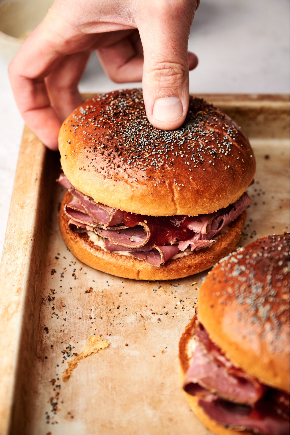 A roast beef sandwich on a baking sheet with a hand placing the top bun on it.