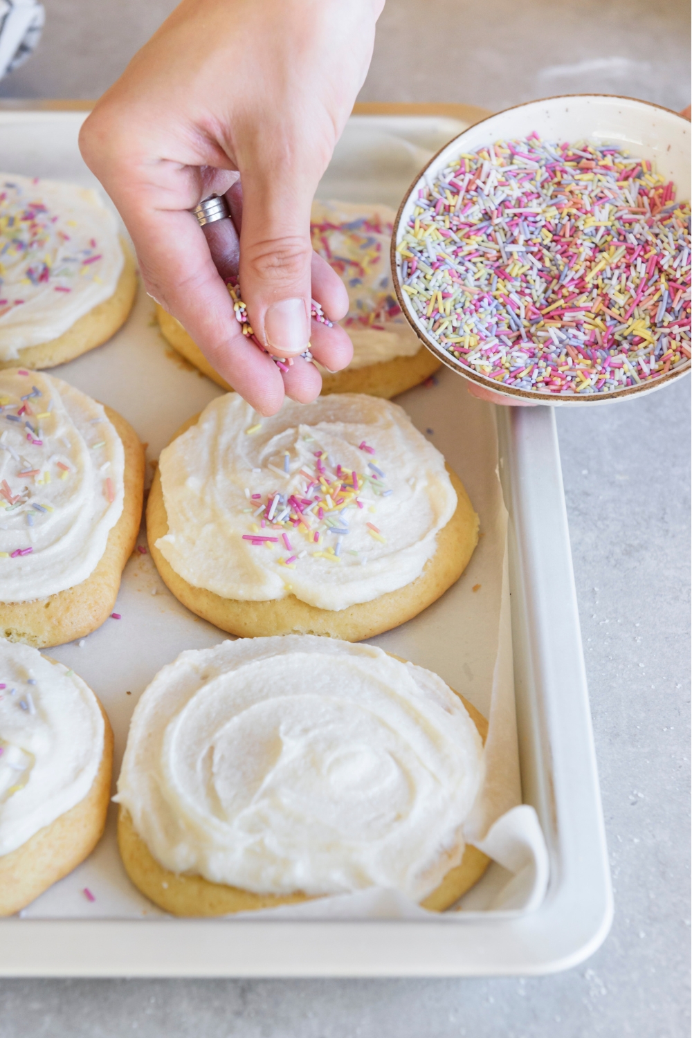 A baking sheet with sugar cookies with frosting. A hand is adding sprinkles to the cookies.