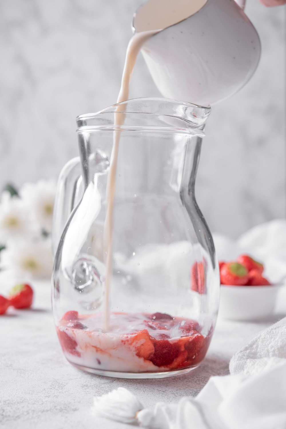 A pitcher with muddled strawberries and a smaller cup with coconut milk being poured into it.