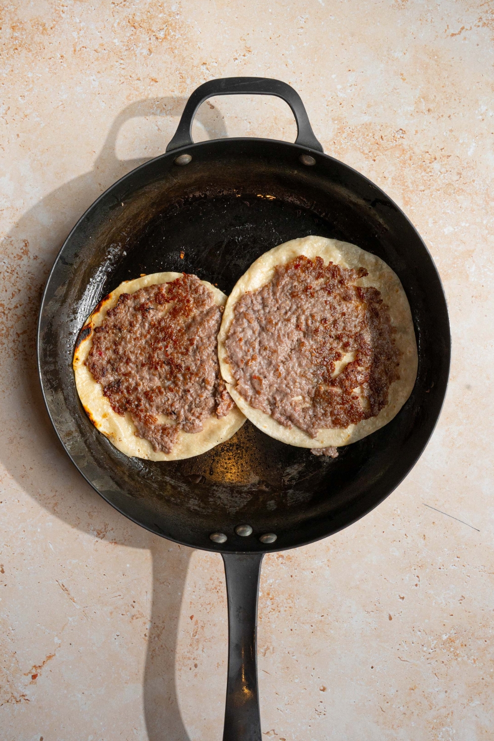 Burger patties on top of tortillas cooking in a black skillet.