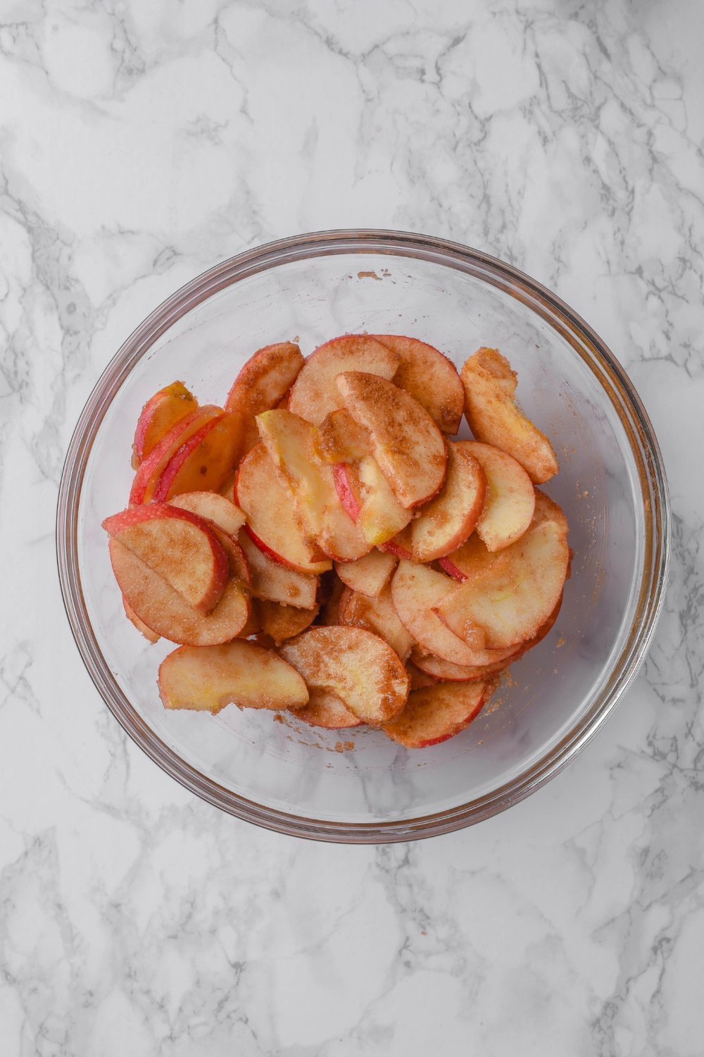 Sliced apples coated in cinnamon sugar in a glass bowl on a grey counter.