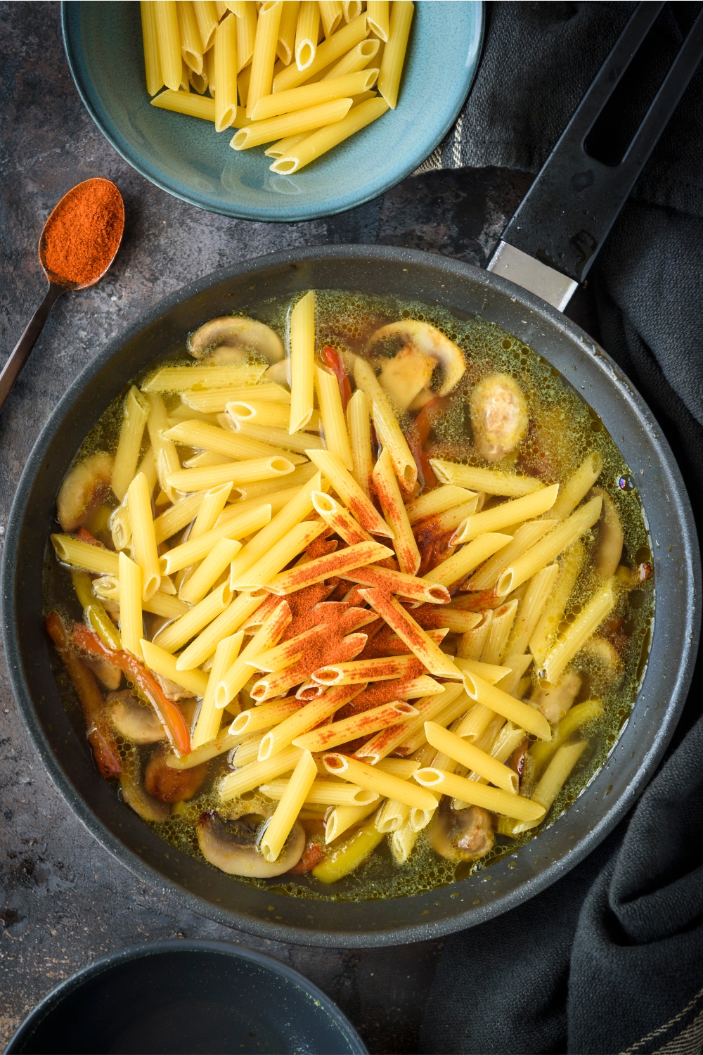 A skillet filled with cooked peppers, cooked mushrooms, and dried pasta with broth and seasonings.