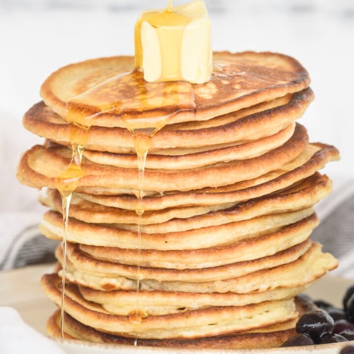 Maple syrup being poured on top of a cube of butter on a stack of ten pancakes on a white plate.
