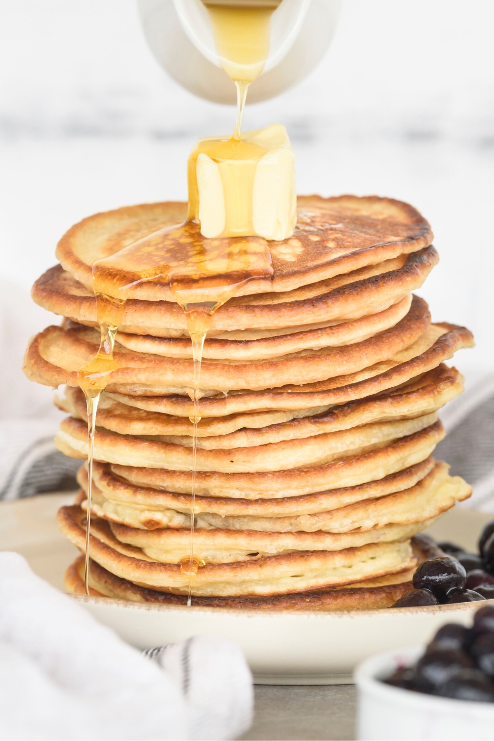 Maple syrup being poured on top of a cube of butter on a stack of ten pancakes on a white plate.