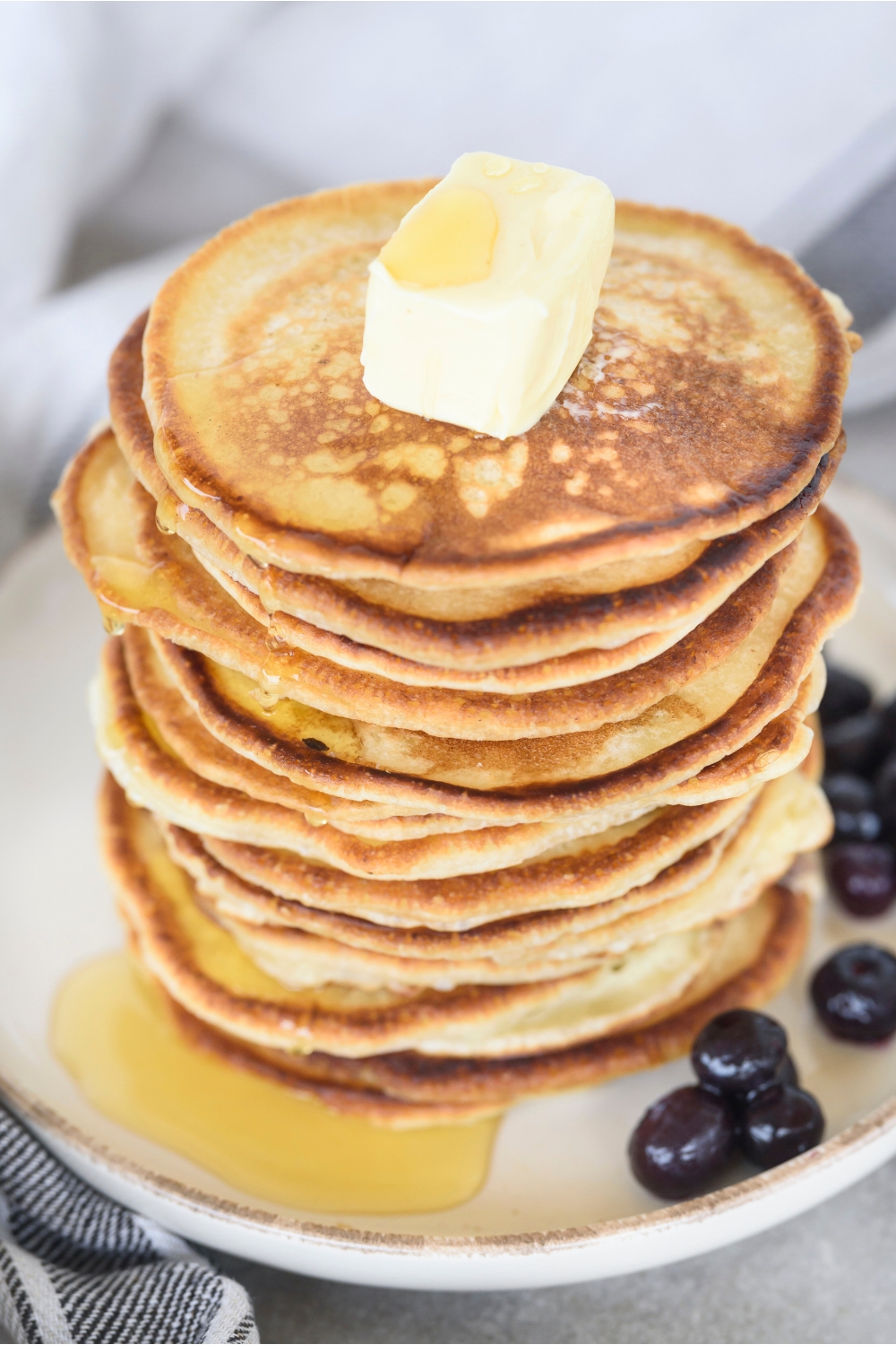 A cube of butter that is on a big stack of pancakes that is on a plat with blueberries.