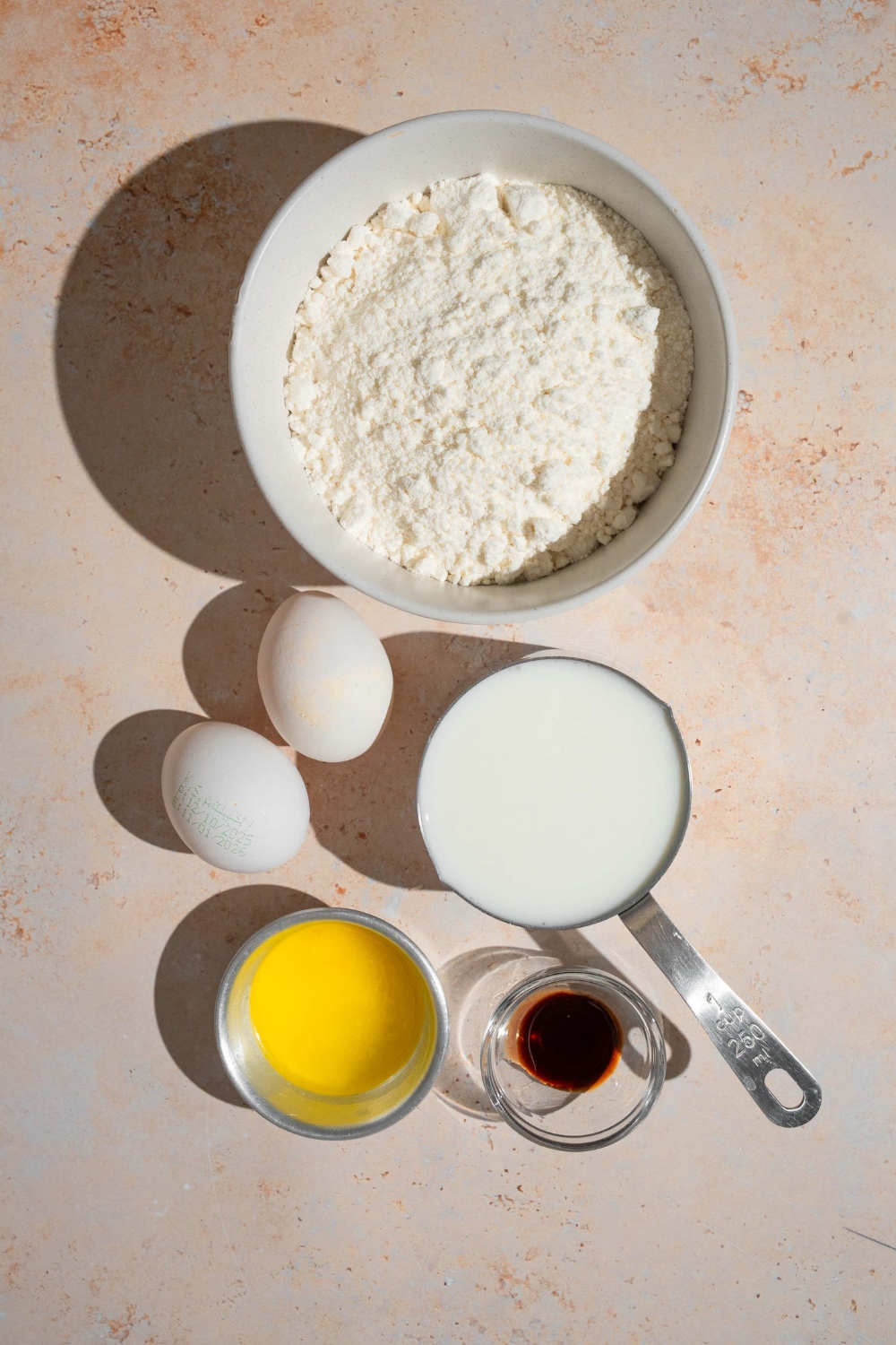 A bowl of flour, two eggs, a cup of milk, a small bowl of butter, and a small bowl of vanilla extract on a white counter.
