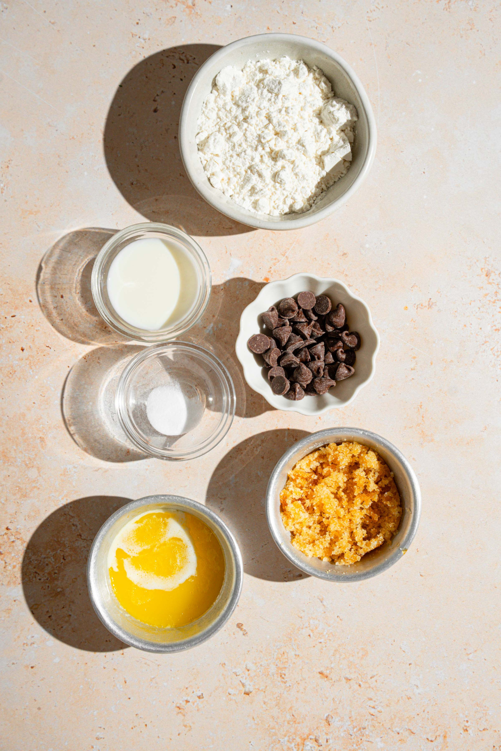 A bowl of flour, a bowl of milk, a bowl of chocolate chip cookies, a bowl of brown sugar, and a bowl of melted butter all on a white counter.