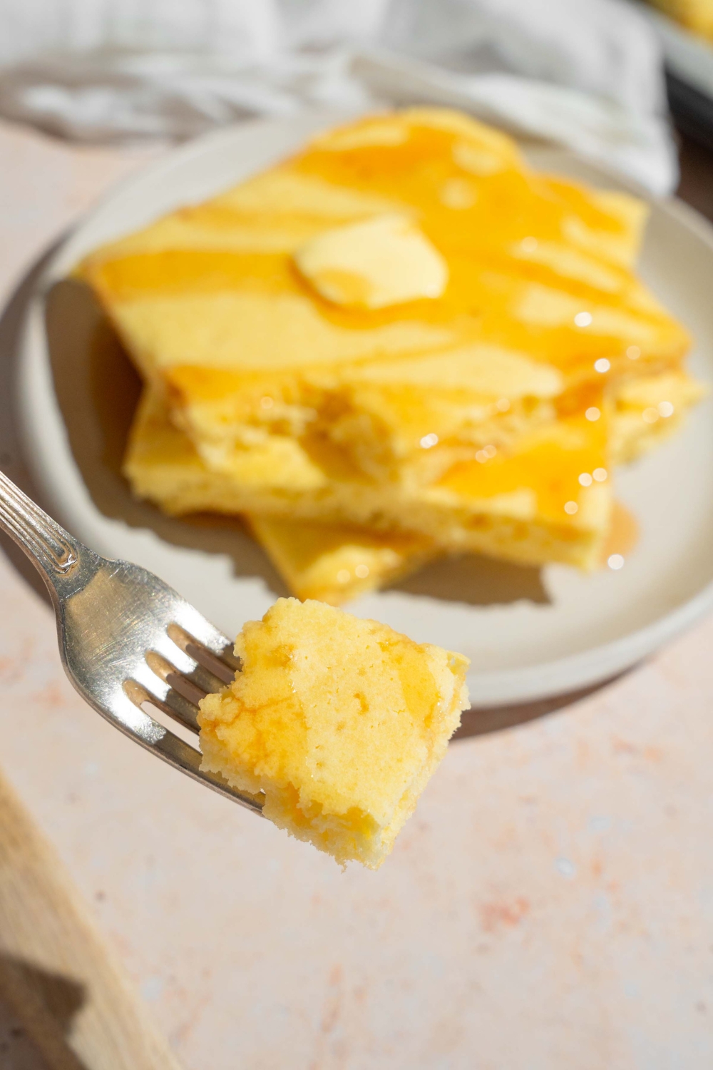 A piece of a sheet pan pancake on a fork that is being held in front of a white plate that has three squares of sheet and pancake stack on top of one another.