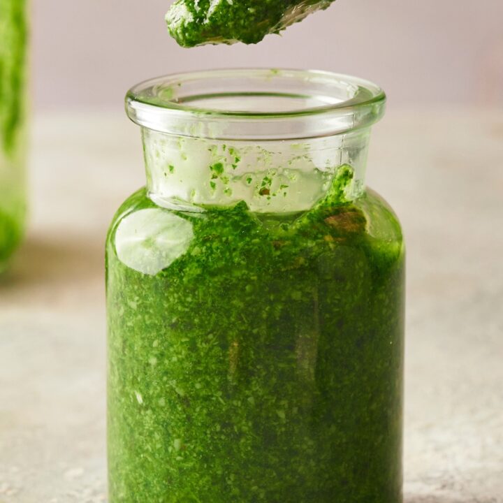 A spoon with some pesto on it above a jar that's filled with pesto on top of a counter.