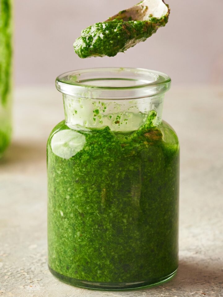 A spoon with some pesto on it above a jar that's filled with pesto on top of a counter.