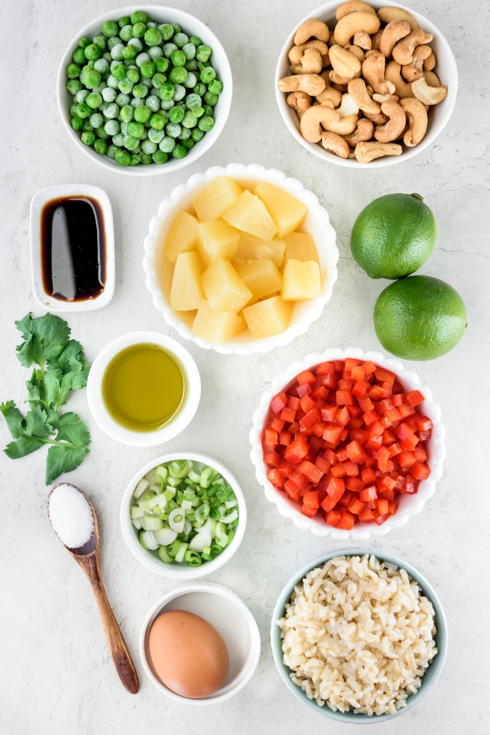 A countertop with pineapple, cashews, peas, soy sauce, olive oil, limes, bell pepper, green onion, rice, salt, cilantro, and an egg in various bowls.