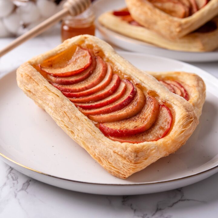 A baked puff pastry dough that has apples overlapping one another down the middle of the dough. This is on top of another puff pastry apple tart on a white plate on a grey counter.