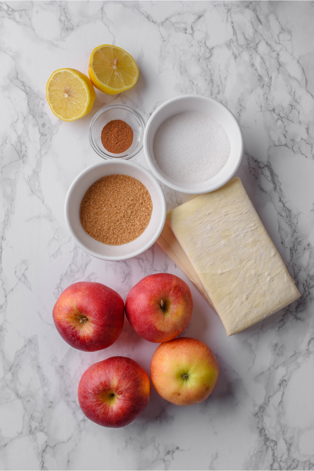 Four apples, a puff pastry block, a halved lemon, and bowls of brown sugar, white sugar, and cinnamon.