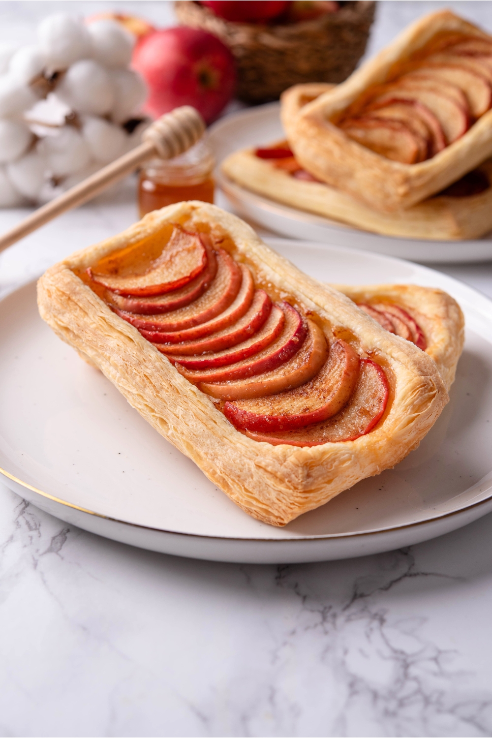 A baked puff pastry dough that has apples overlapping one another down the middle of the dough. This is on top of another puff pastry apple tart on a white plate on a grey counter.