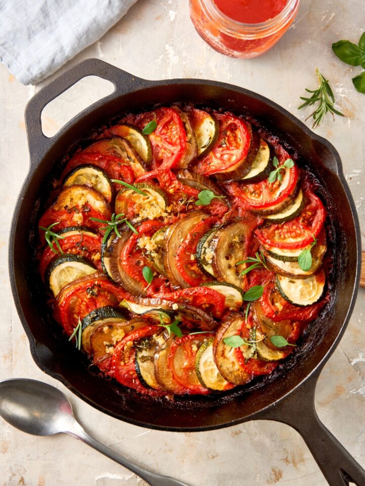 Tomatoes, zucchini, and eggplant slices arranged in an overlapping circle in a cast iron skillet on top of a white counter.