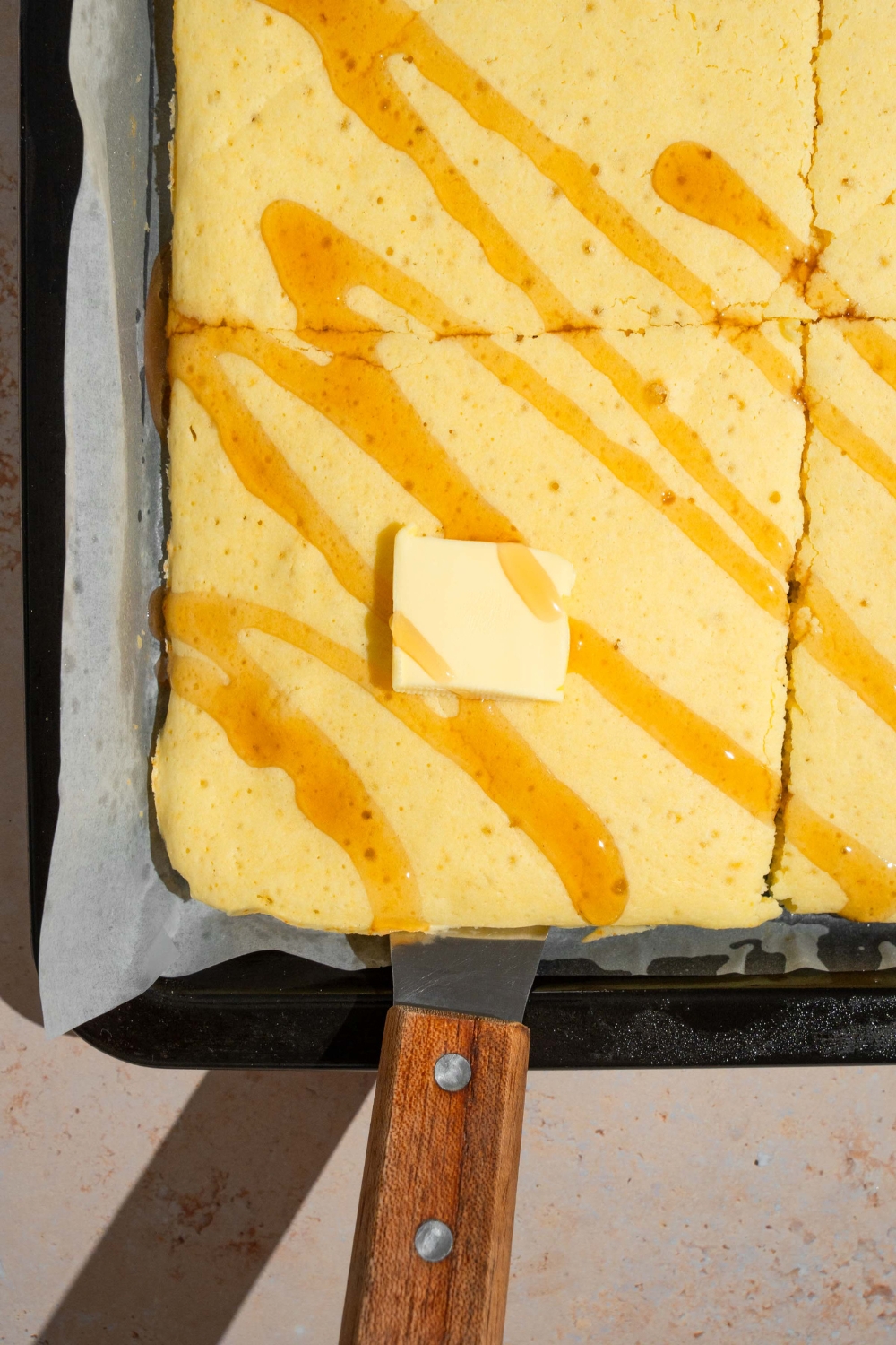Butter square on top of a square of a sheet pan, pancake with other squares of sheet pan, pancakes around it on a sheet of parchment paper and a baking sheet.