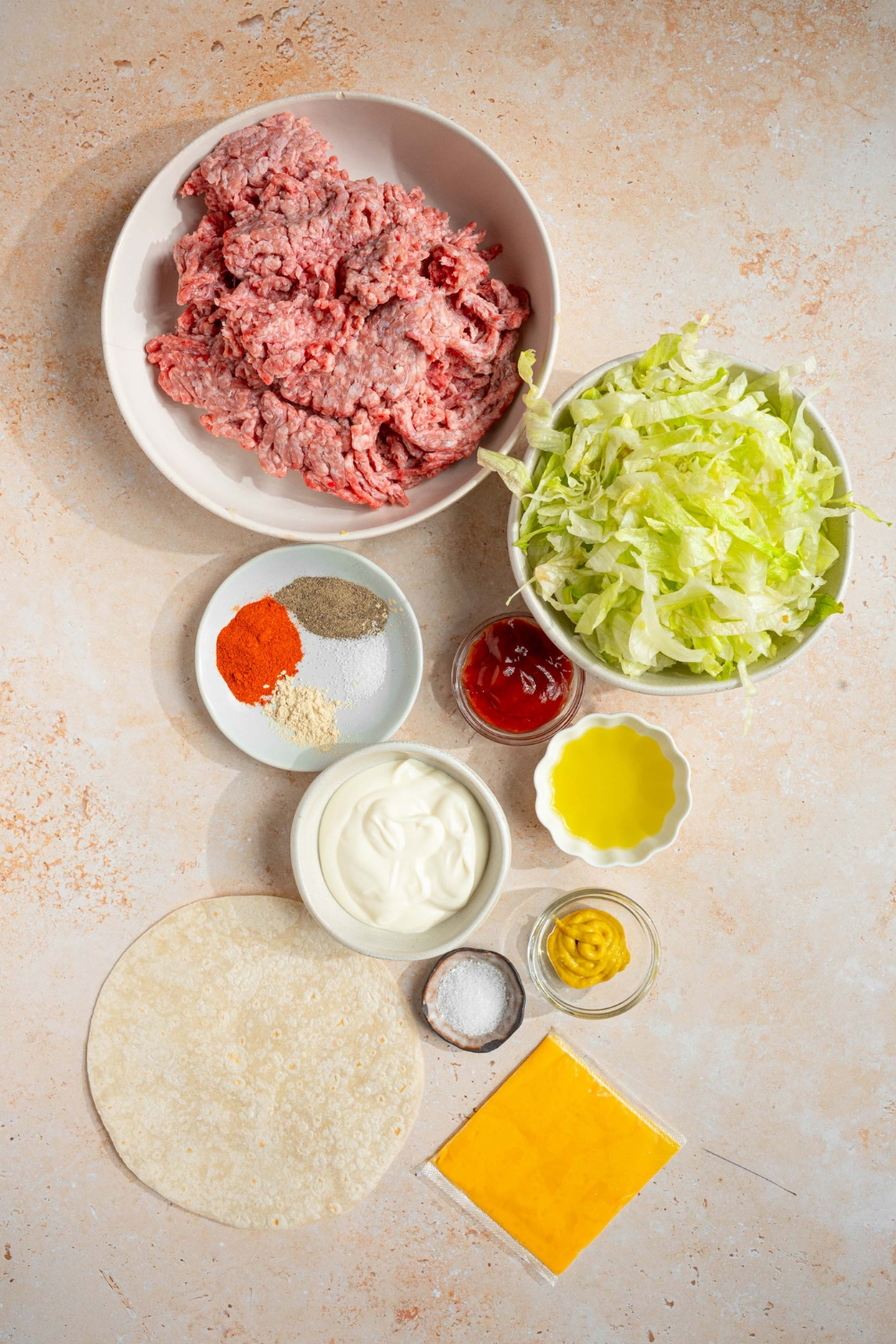 A bowl of ground beef, a bowl of lettuce, a bowl of ketchup, a bowl of mustard, a bowl of salt and pepper, a bowl of mayo, tortillas, and slices of cheddar cheese all on a white counter.