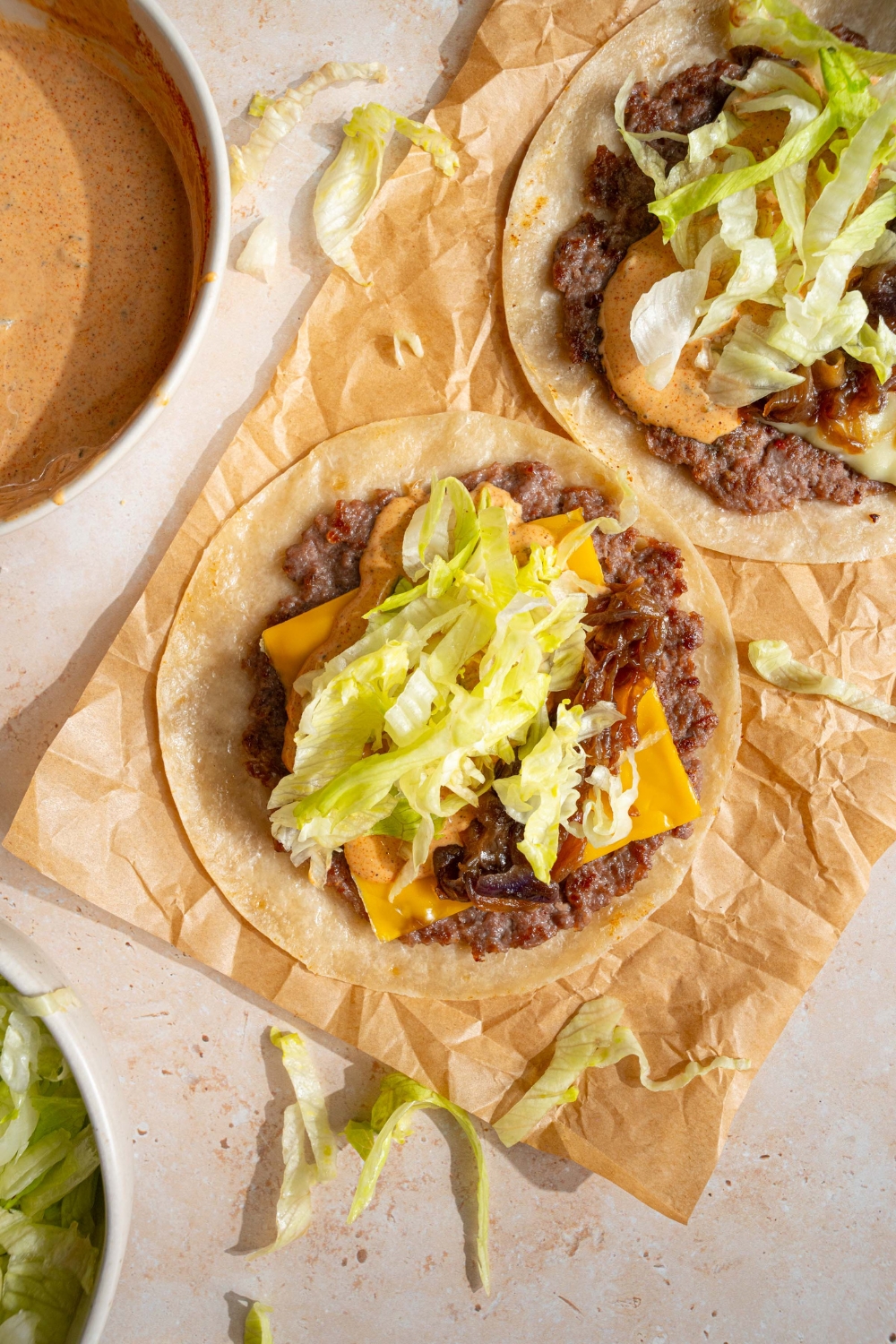Shredded lettuce on top of cheese and a smash burger patty on a tortilla on a piece of parchment paper.