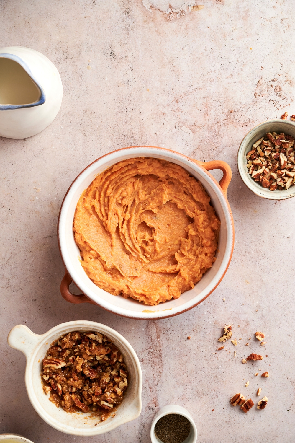 A white pot full of mashed sweet potatoes. A mixing bowl pull of pecan topping stands nearby.